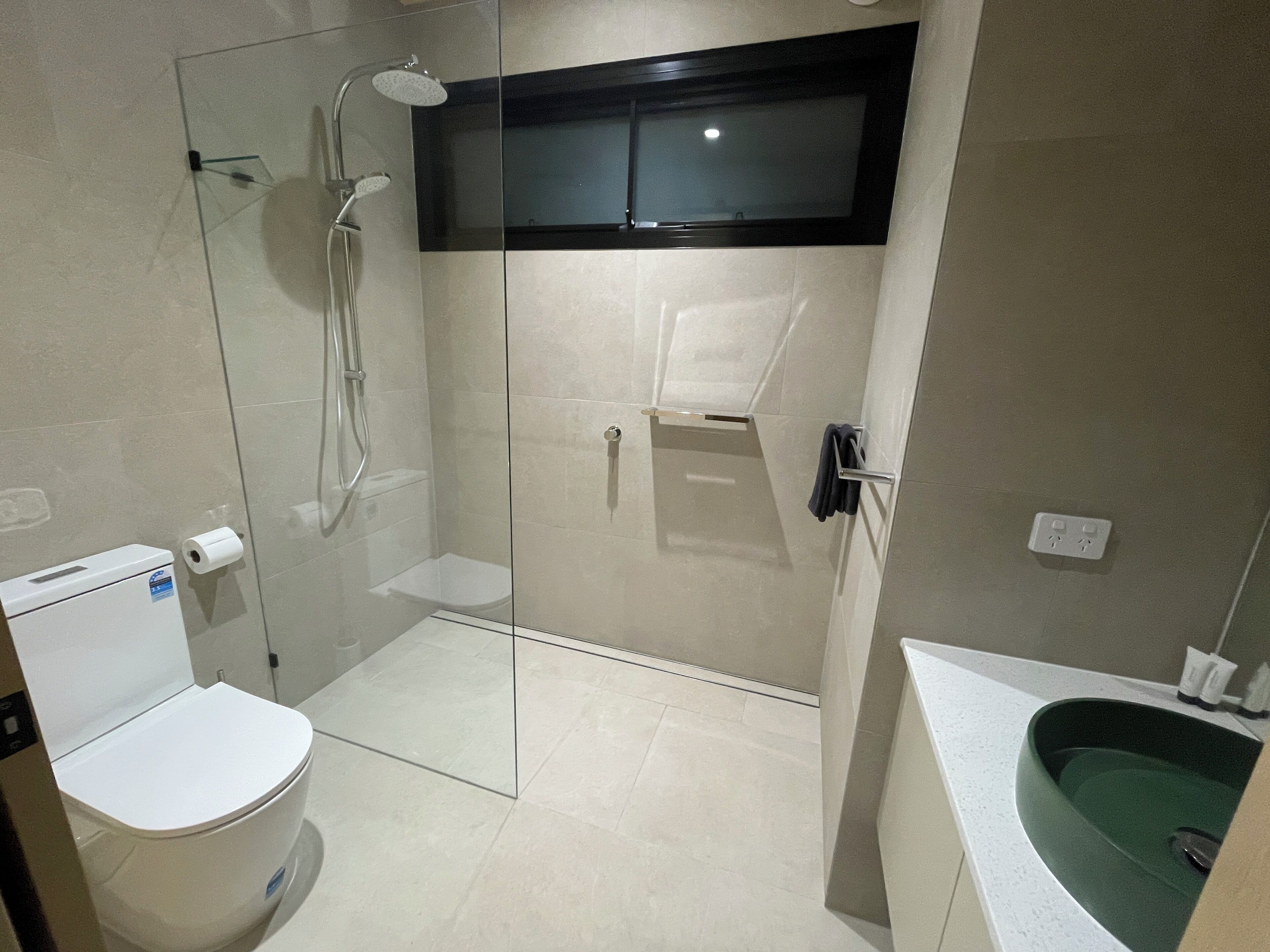 Un baño moderno con ducha acristalada, inodoro, lavamanos de color verde y un perchero para toallas.