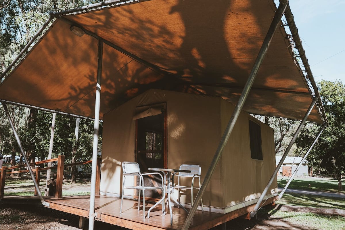 A tent with a porch and two chairs, set in a wooded area with a vehicle in the background.
