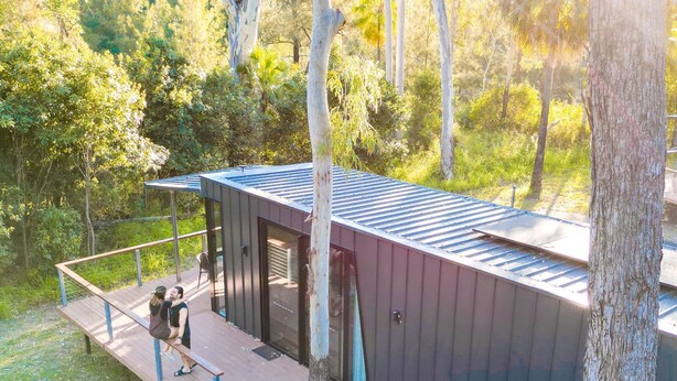 Una cabaña pequeña y moderna con un balcón y un espacio exterior cubierto, rodeada de árboles.