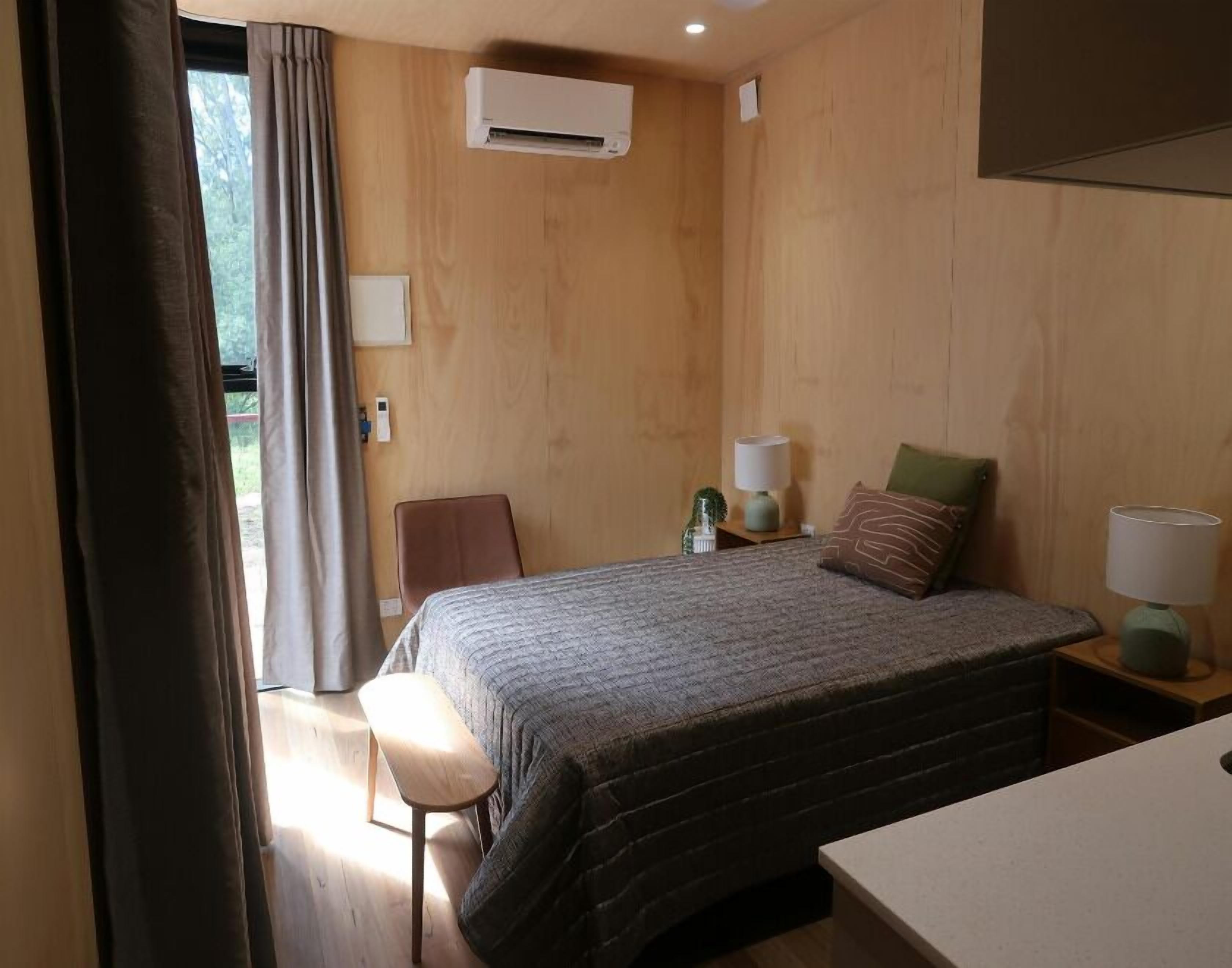 A hotel room with a bed, bedside tables, a chair, an air conditioning unit, and a window with curtains.