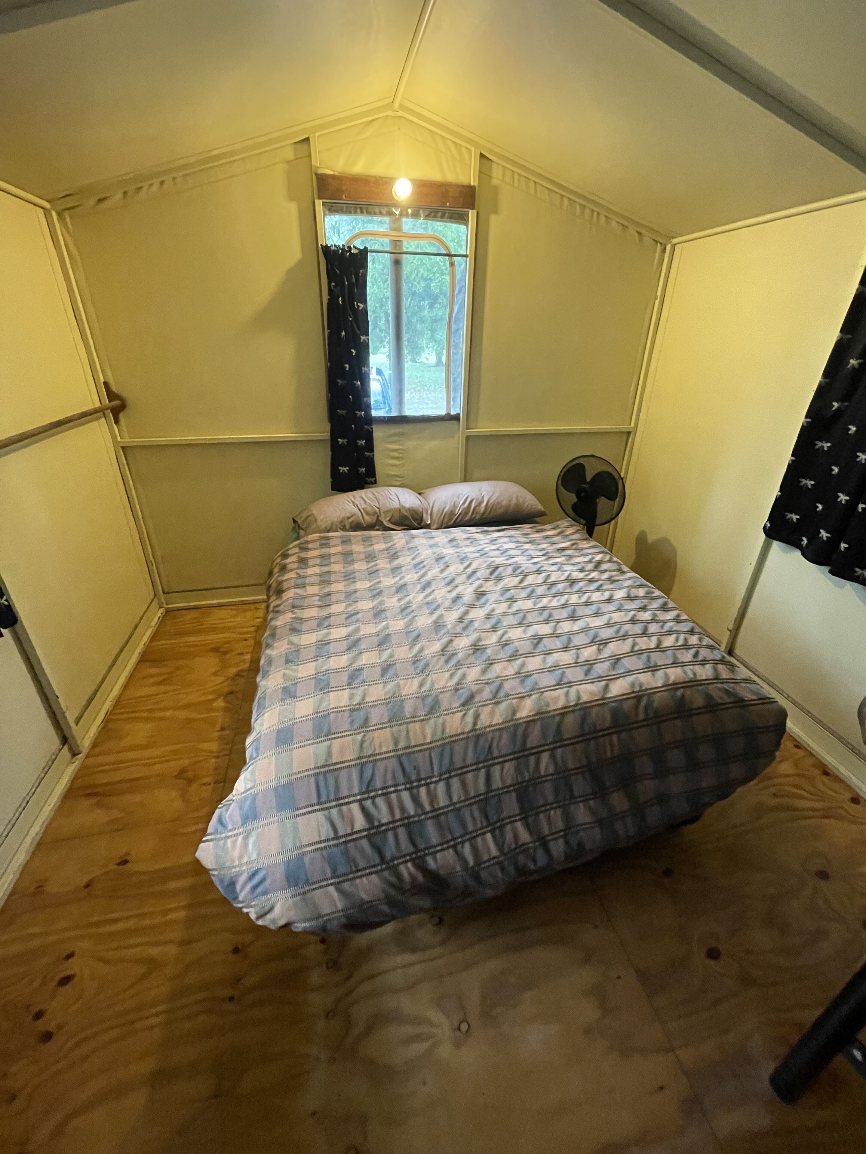 Une petite pièce agréable, dotée d’un lit simple, d’une fenêtre avec des rideaux et d’un ventilateur de plafond.