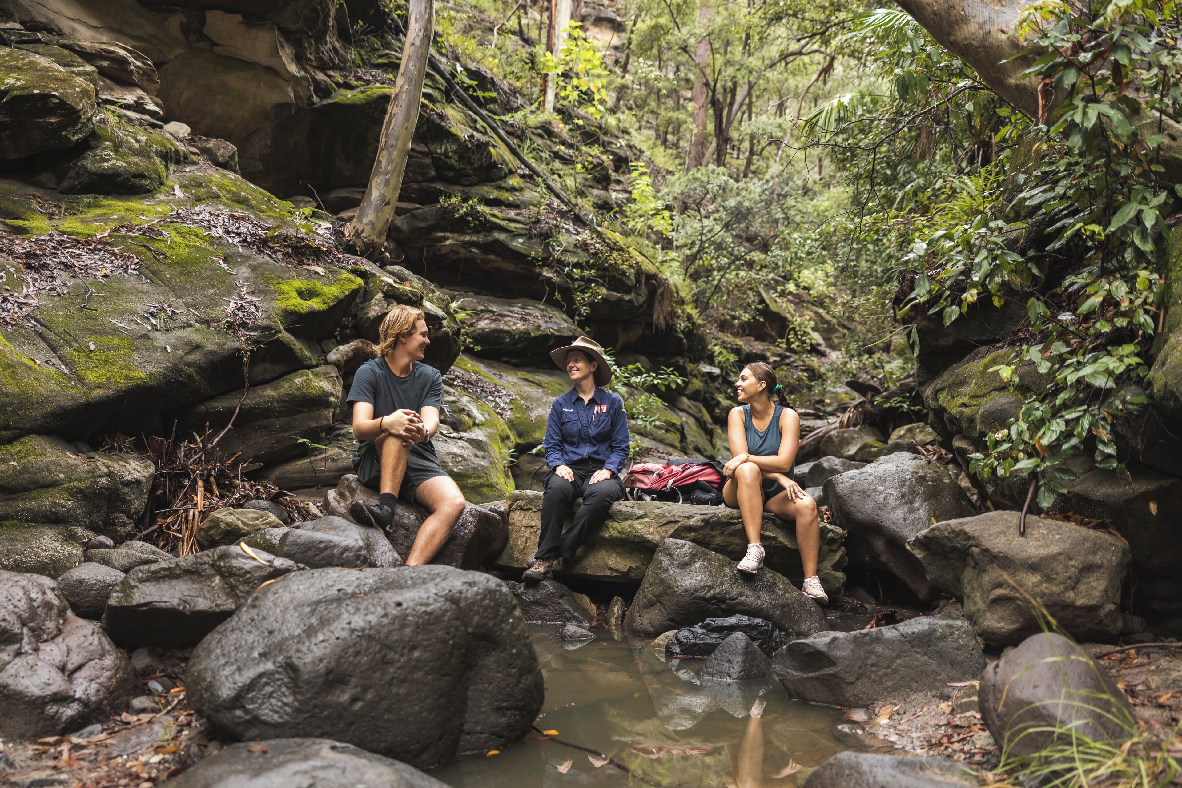 Three people sitting on rocks by a stream in a forested area.