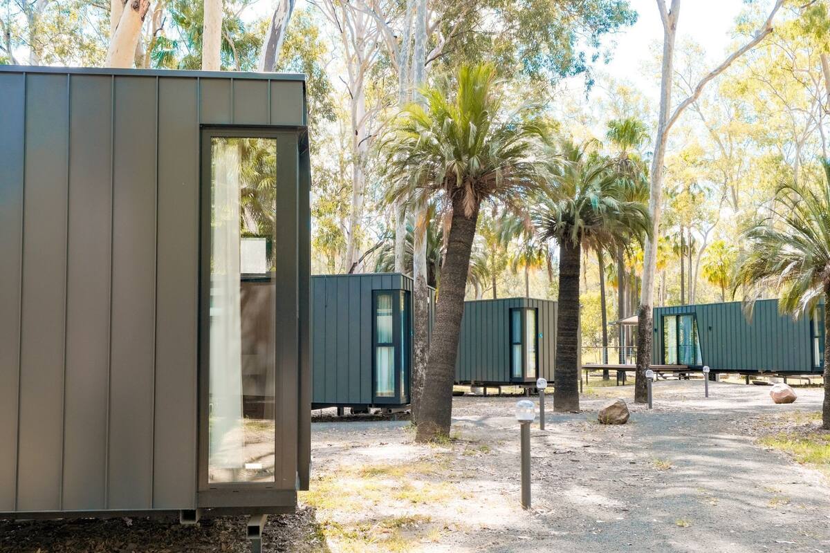 A row of modern, compact cabins with a natural backdrop of trees and a dirt ground.
