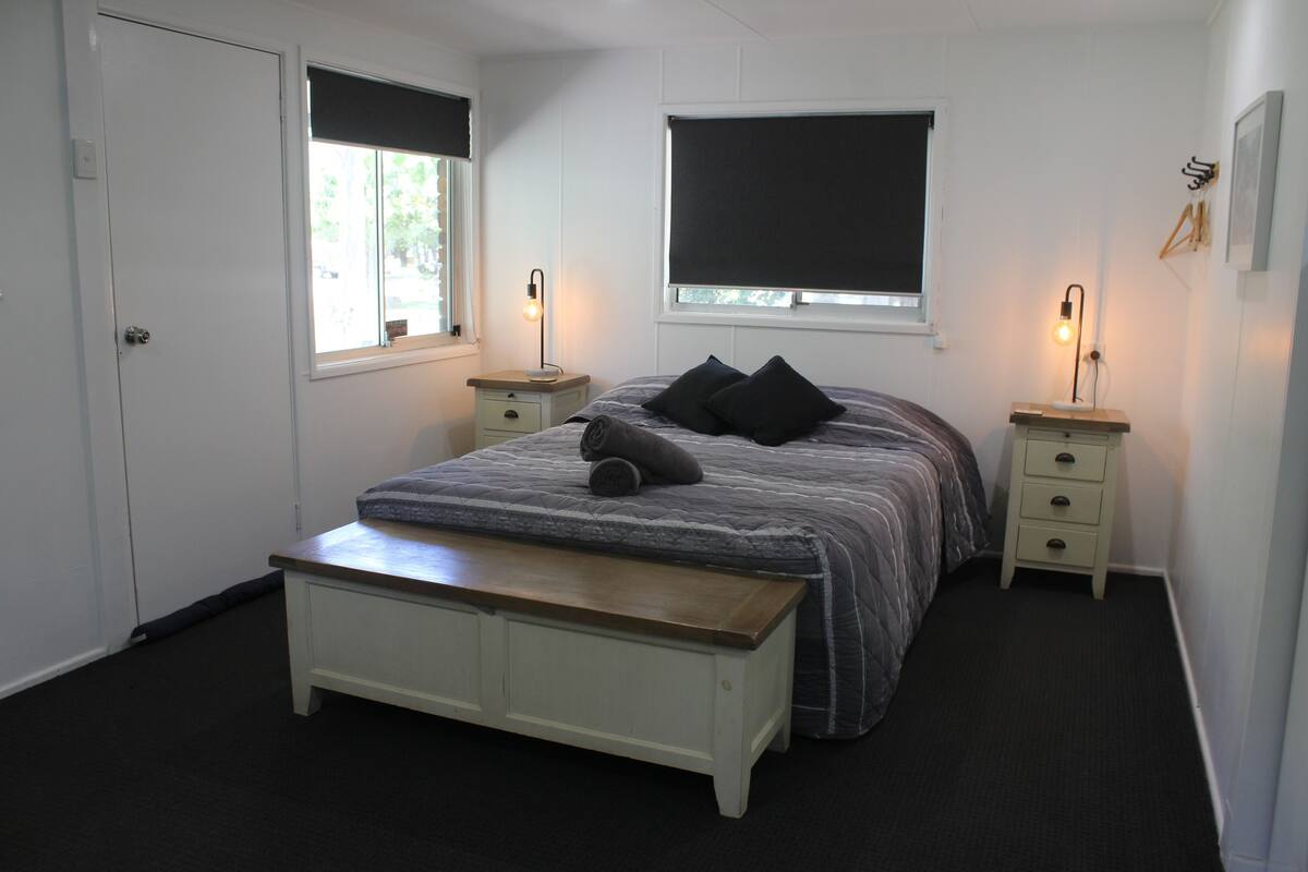 A bedroom with a bed, two bedside tables, a bench, a window with blinds, and a door.