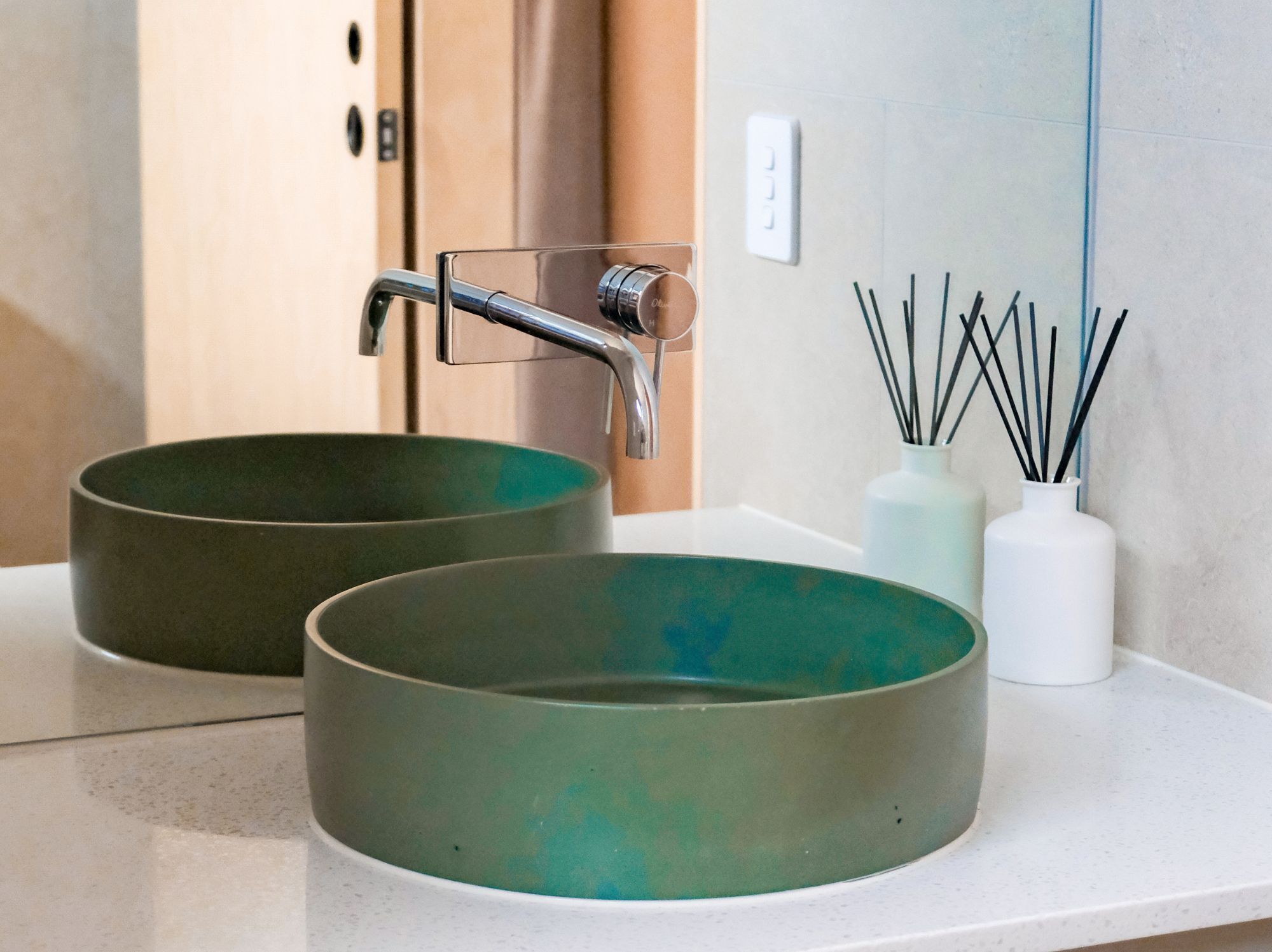 A bathroom with two round green sinks, a modern faucet, and a white vase with reed sticks.