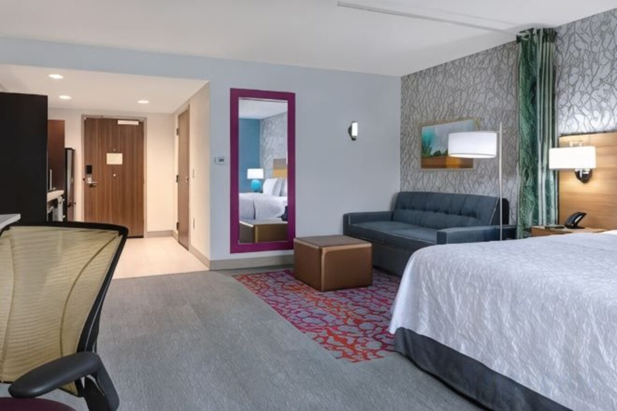 Habitación de hotel con cama, sofá, escritorio, silla, espejo y puerta que da a otra habitación.