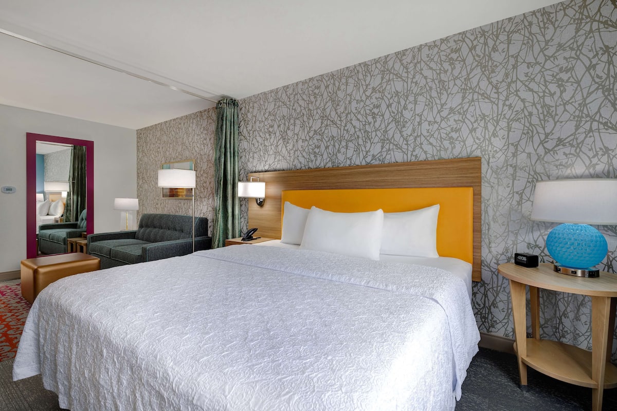 A hotel room with a large bed, a nightstand, a chair, a desk, and a mirror.