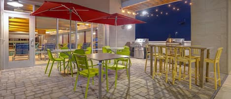 Ein Außenbereich mit einer Pflasterterrasse, grünen und gelben Stühlen, einem roten Sonnenschirm und einem Grill.