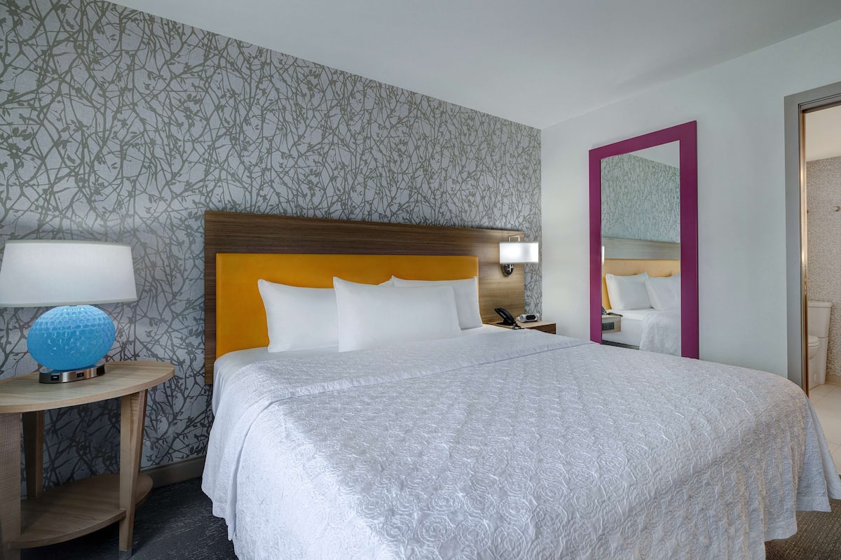 A hotel room with a large bed, a nightstand, a lamp, and a mirror.
