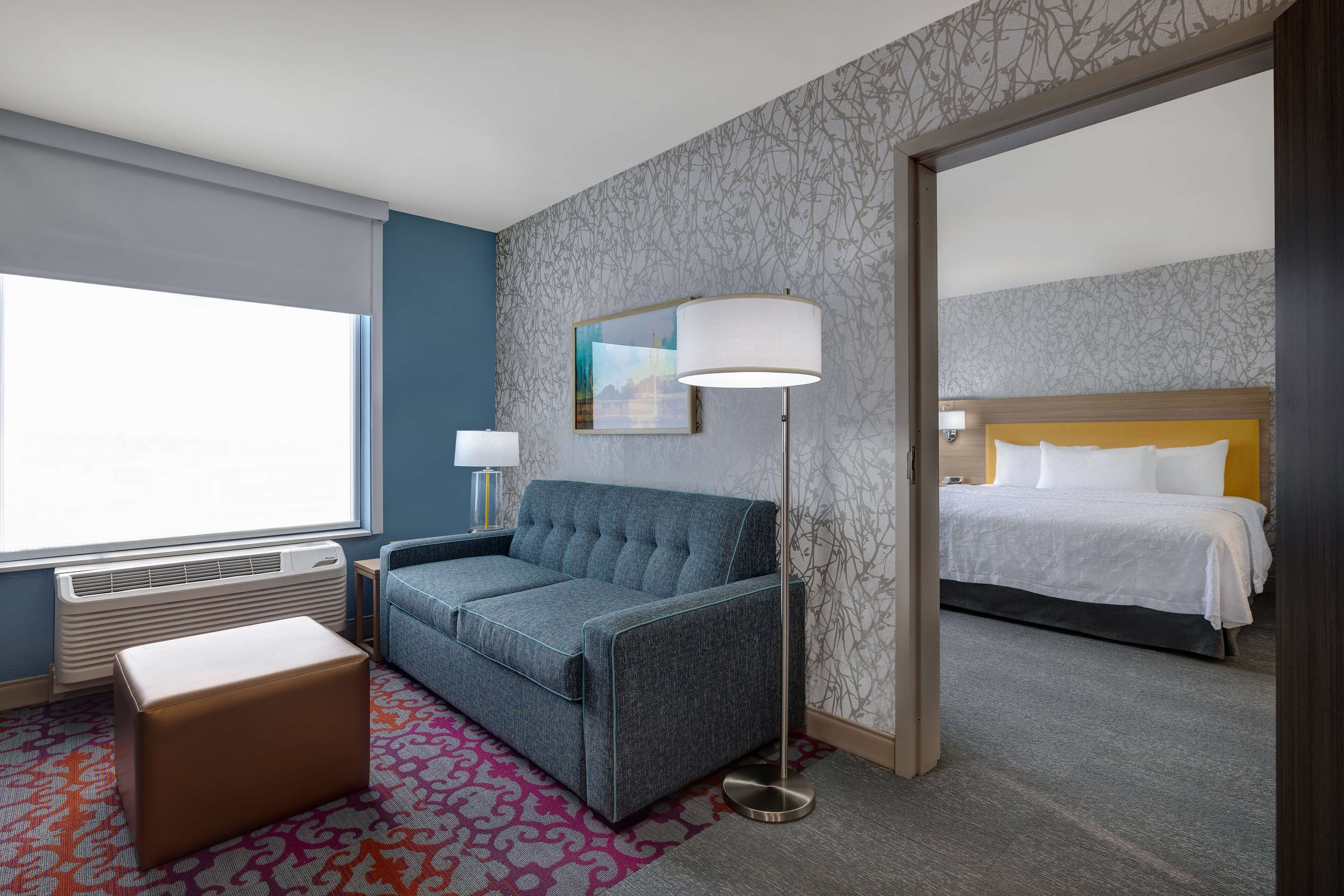 A hotel room with a sofa, a bedside table, a lamp, a patterned rug, and a bed with a headboard.