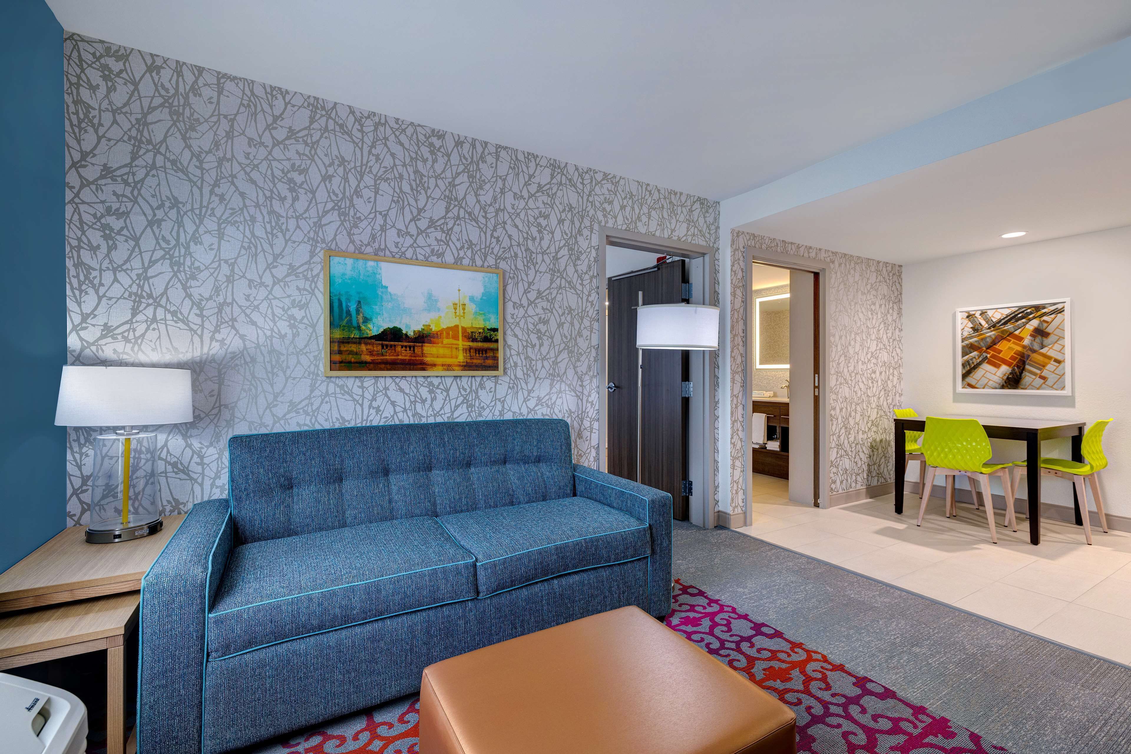 Uma sala de estar moderna com um sofá azul, uma mesa lateral de madeira, uma luminária, um tapete estampado e uma área de refeições com mesa e cadeiras.