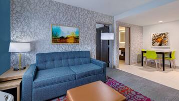 Uma sala de estar moderna com um sofá azul, uma mesa lateral de madeira, uma luminária, um tapete estampado e uma área de refeições com mesa e cadeiras.