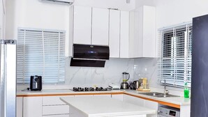 Une cuisine moderne avec des meubles blancs, un îlot central, un lave-linge encastré et une fenêtre avec des stores.