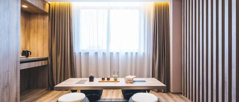 A modern hotel room with a wooden floor, a large window with sheer curtains, a wooden table with a tray of items, and a seating area with two stools.