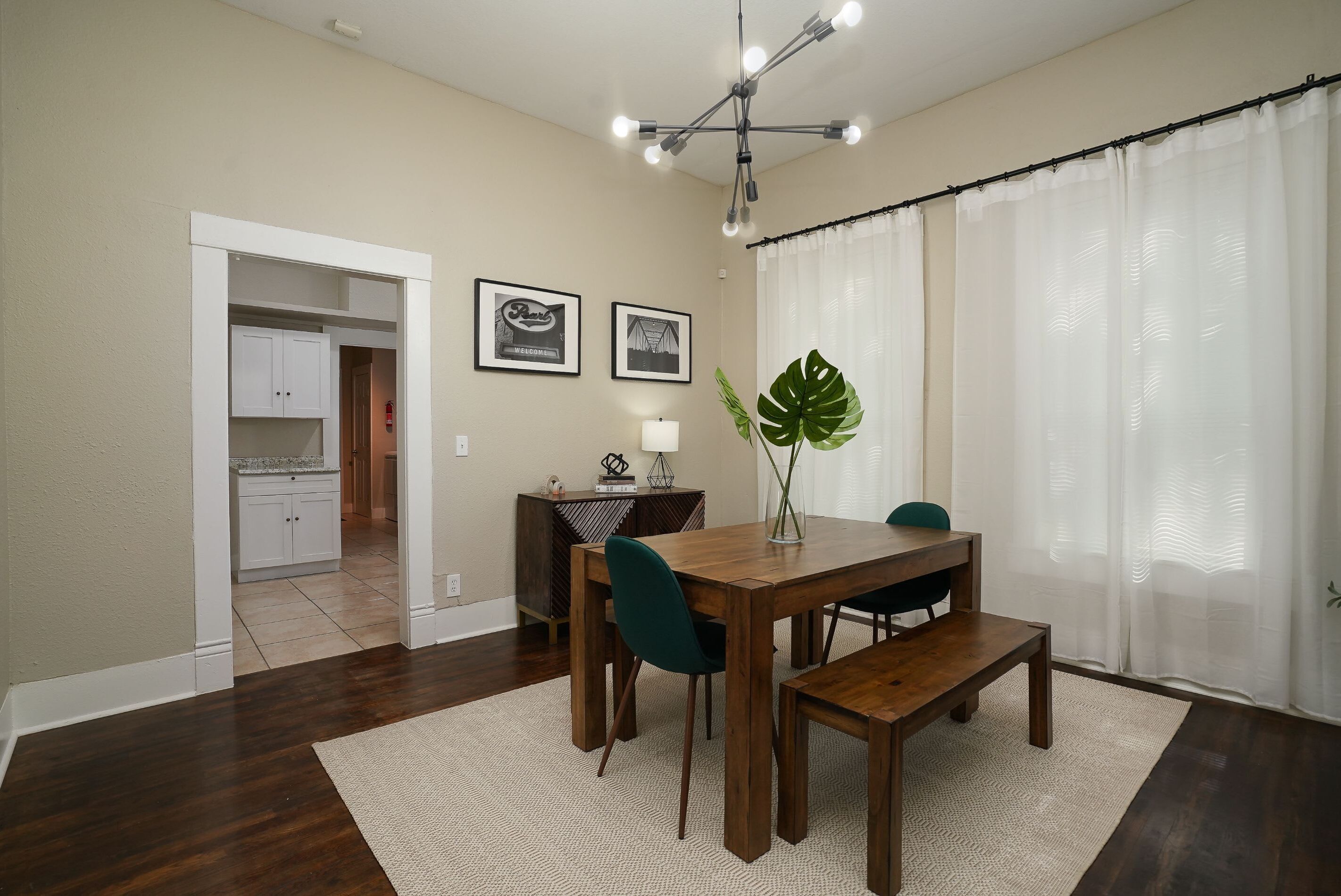 Un coin repas avec une table en bois, des chaises et un banc. On aperçoit une plante sur la table et une lampe sur une petite table d’appoint. La pièce a un plafond et des murs blancs, et une porte qui mène à une autre pièce.