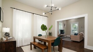 Een eetgedeelte met een houten tafel, stoelen en een bank. Op tafel staat een grote plant, en er is een televisie op de achtergrond.