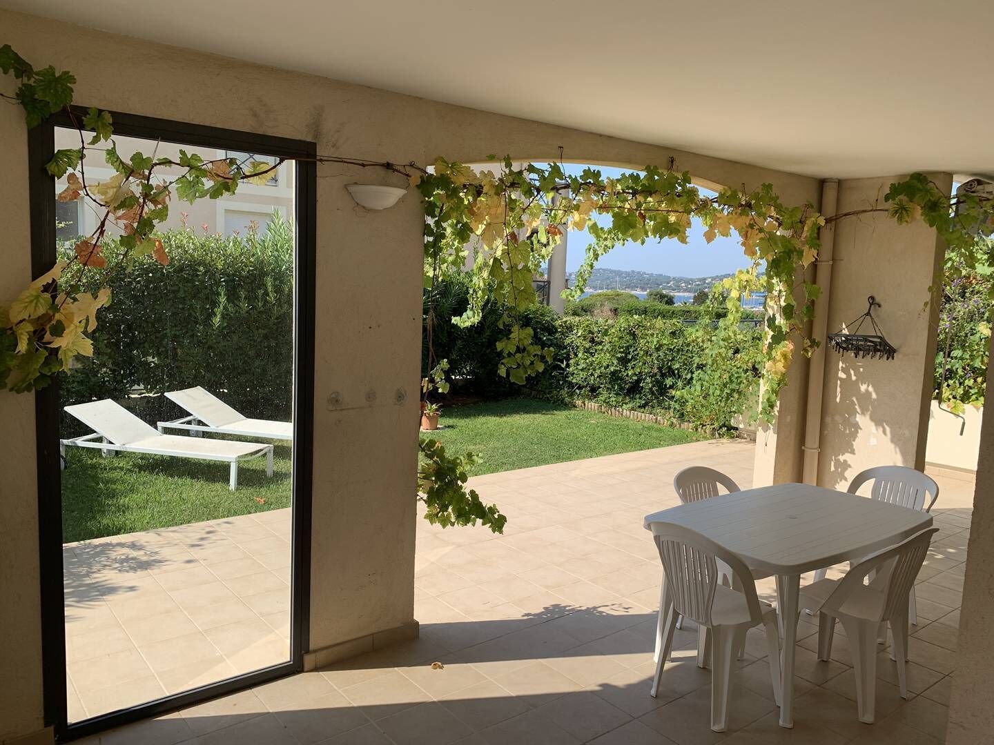 Une terrasse avec une table et des chaises, une porte vitrée donnant sur un jardin aménagé avec des fauteuils de jardin.