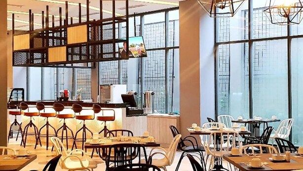 Restaurante com pé-direito alto, amplas janelas e luminárias geométricas modernas.