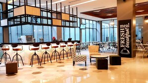 Uma área de café da manhã moderna com mesas e cadeiras altas, um balcão com cardápio e grandes janelas com vista para a cidade.