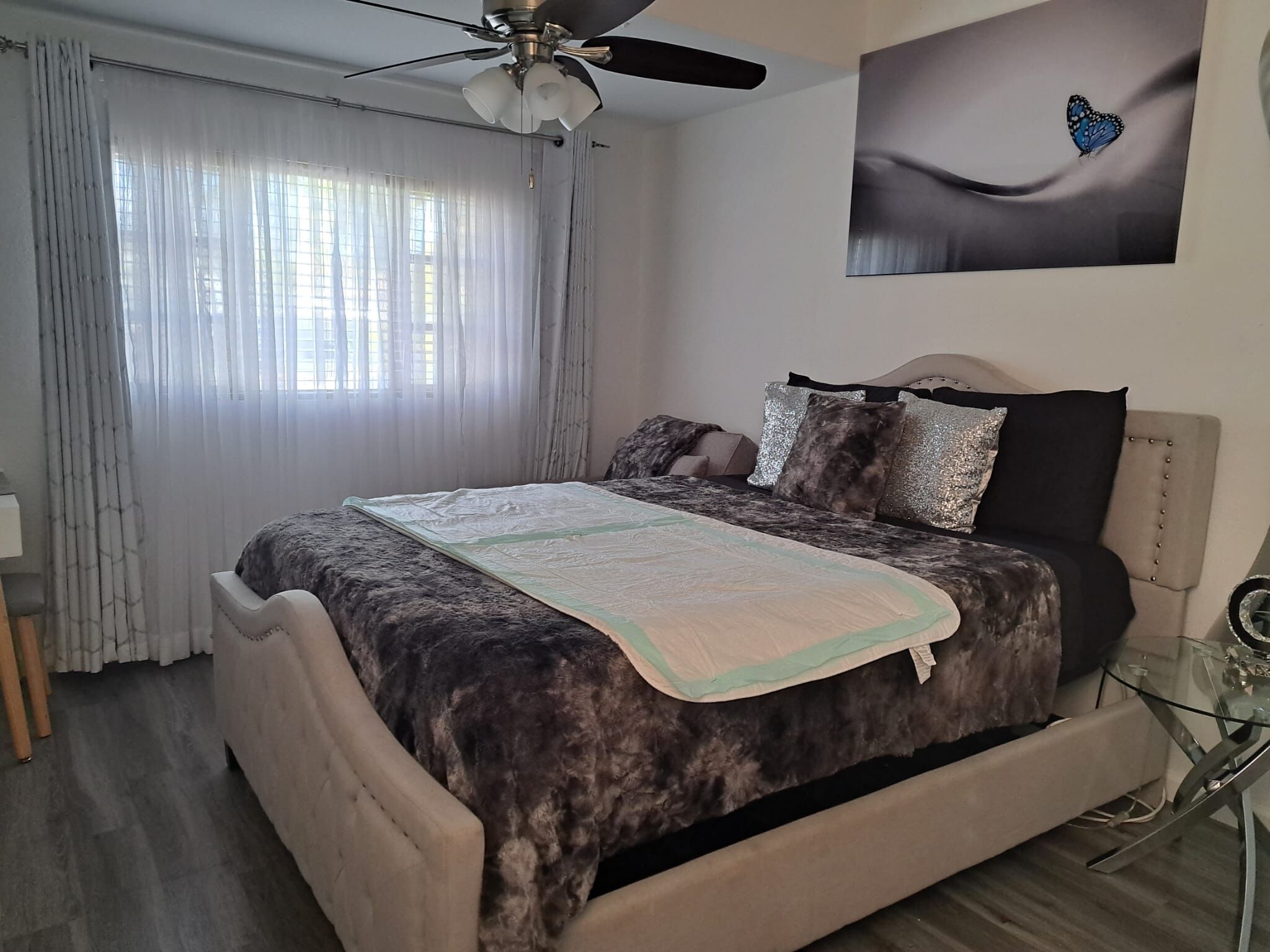 Un dormitorio con una cama, ventilador de techo, ventana con cortinas e imagen colgada en la pared.