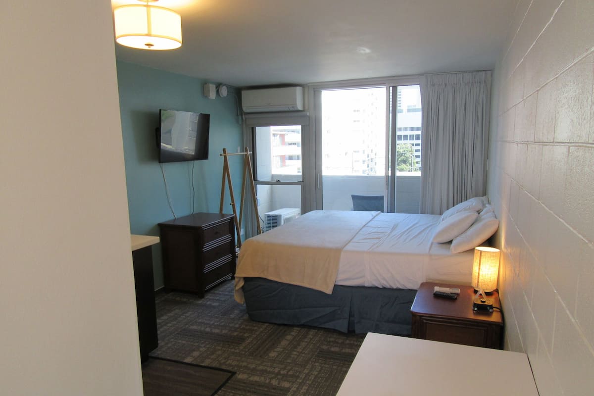 Habitación de hotel con cama, escritorio, cómoda, televisor y ventana con cortinas.
