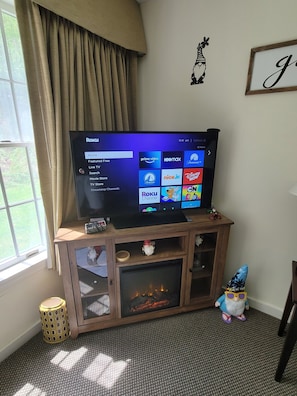 A television on a wooden stand with a fire feature below it, a small gnome figurine, and a framed picture on the wall.