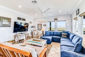 A living room with a large flat-screen TV, a blue sectional sofa, a wooden coffee table, and a ceiling fan.