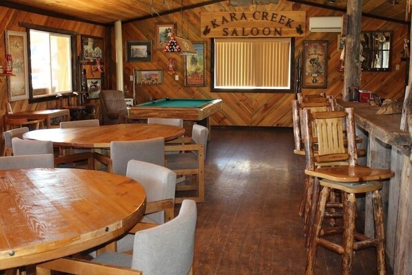 Un coin bar rustique avec des tables et des chaises en bois, une table de billard et une enseigne indiquant « KARA CREEK SALOON ».