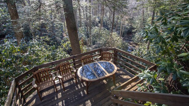 Une terrasse en bois avec une table et des chaises à plateau en verre, le tout entouré d’une forêt dense.