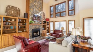 Un salon chaleureux avec une cheminée en pierre, des étagères en bois, des fauteuils en cuir et de grandes fenêtres.