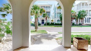 A view through a covered outdoor area with a paved walkway, palm trees, and a street in the background. - Designer Condo in Popular Palmilla | Close to Pool & Boardwalk (Port Aransas)