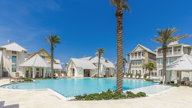 A pool area with a clear blue sky, surrounded by multi-story buildings with balconies and palm trees. - Designer Condo in Popular Palmilla | Close to Pool & Boardwalk (Port Aransas)