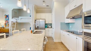 A modern kitchen with white cabinets, a central island with a sink, stainless steel appliances, and pendant lighting. - Designer Condo in Popular Palmilla | Close to Pool & Boardwalk (Port Aransas)