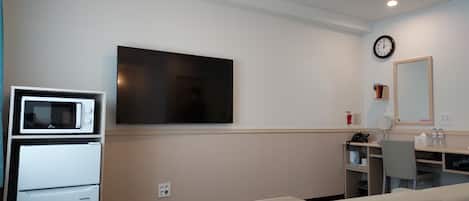 A modern hotel room with a flat-screen TV, a microwave, a refrigerator, a sofa, a desk with a chair, a clock, and a mirror.