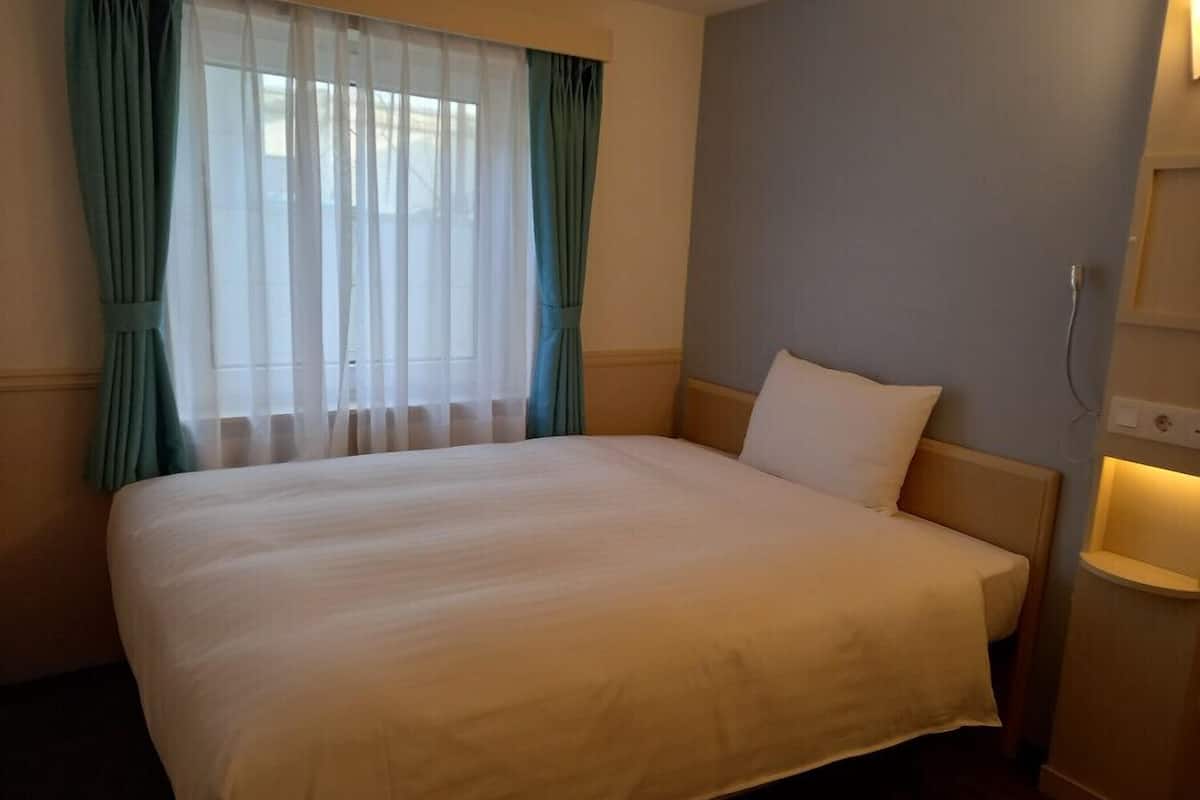 A hotel room with a large bed, a window with sheer curtains, a bedside table with a lamp, and a wall-mounted light.