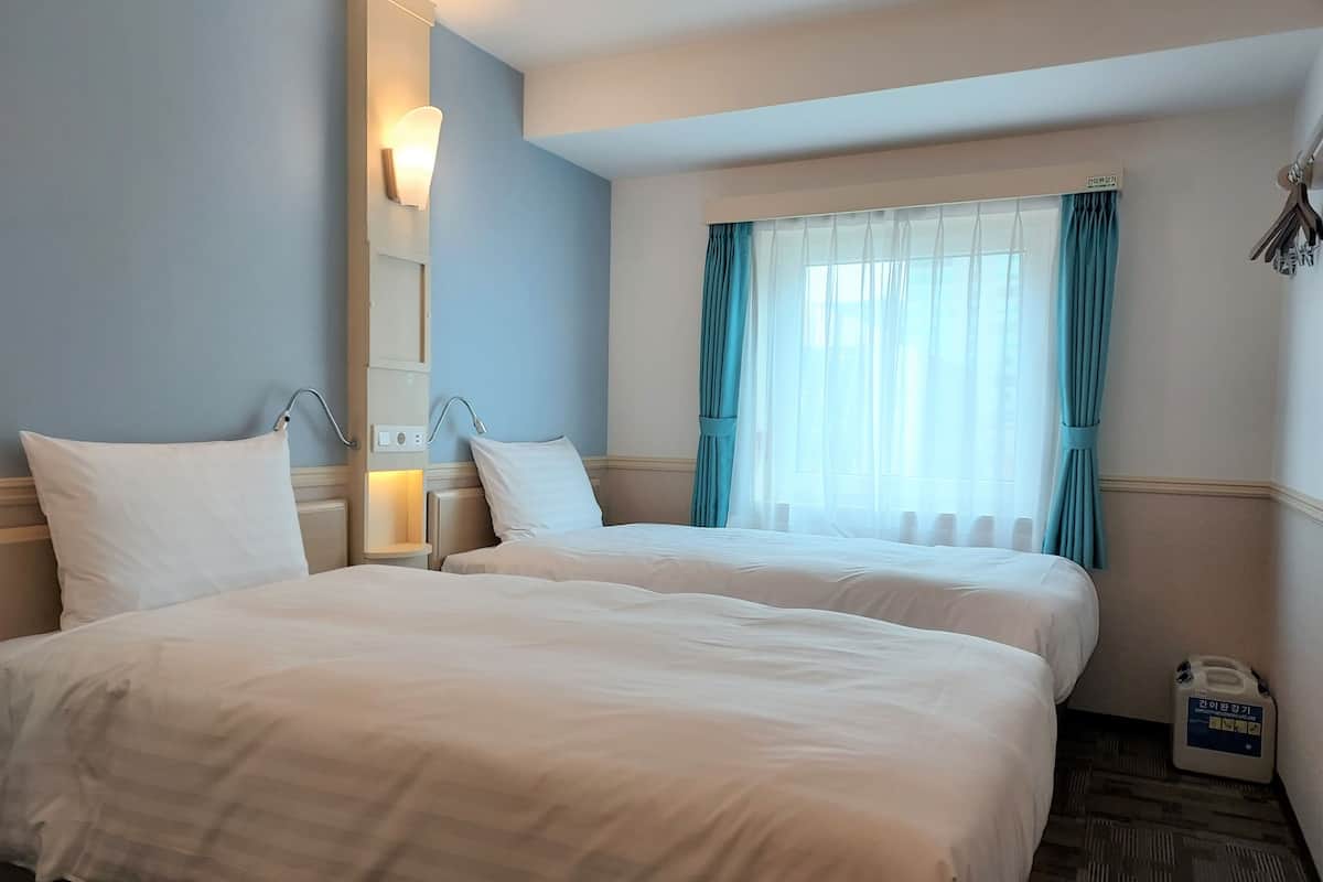 A hotel room with two beds, a window with curtains, a wall-mounted light, and a small table with a bottle.