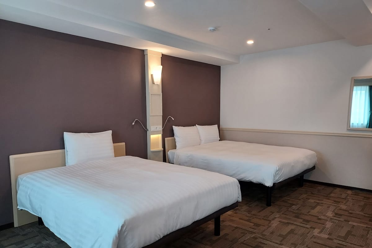 A hotel room with two single beds, a mirror, a wall-mounted light, and a window with curtains.