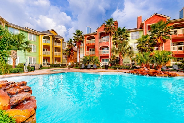 A swimming pool with a waterfall feature, surrounded by colorful buildings and palm trees.