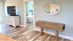 Een kamer met een houten console, een televisie op een wit kastje, een ronde spiegel aan de muur, en een deur naar een slaapkamer.