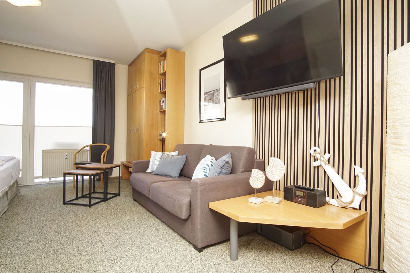 A hotel room with a sofa, a small table, a chair, a TV, a bookshelf, and a radiator.