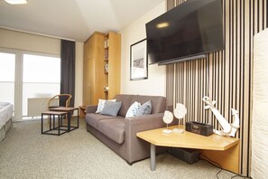 A hotel room with a sofa, a small table, a chair, a TV, a bookshelf, and a radiator. - Just 100 m from the beach, apartment with a balcony - Apt. Eldena FeWo 15 - Apartments (Middelhagen)