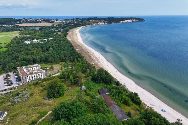 An aerial view of a coastal area with a beach, a large building with multiple floors, and a parking lot.