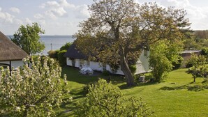 A coastal house with a large tree in the front yard, a thatched-roof building to the left, and a view of the sea in the background. - Beachfront idyll by the sea, private bathing area - Gutshof Ostseeblick FeWo 02 - (Putbus)