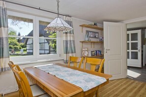 A dining area with a wooden table, chairs, a lace table runner, a hanging light fixture, and a view of a traditional house through the windows.
