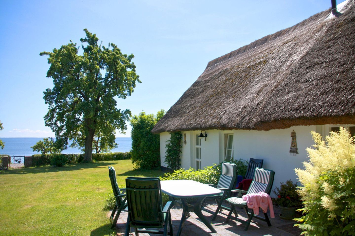 A thatched-roof cottage with a table and chairs in the garden, overlooking a body of water.