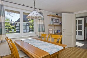 A dining area with a wooden table, chairs, a lace table runner, a hanging light fixture, and a view of a traditional house through the windows. - A Seaside Getaway, Private Swimming Area - Gutshof Ostseeblick FeWo 02 - (Putbus)
