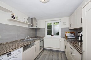 A modern kitchen with grey tiles, a built-in oven, a washing machine, and a window with a view of greenery.