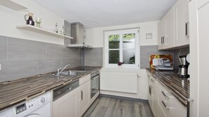 A modern kitchen with grey tiles, a built-in oven, a washing machine, and a window with a view of greenery. - Beachfront idyll by the sea, private bathing area - Gutshof Ostseeblick FeWo 02 - (Putbus)