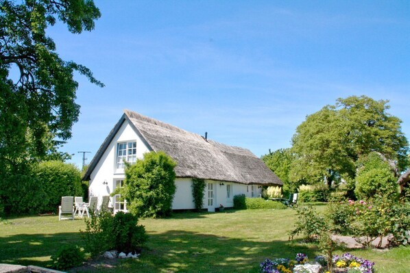 A white house with a thatched roof, surrounded by a well-maintained garden with various plants and flowers.