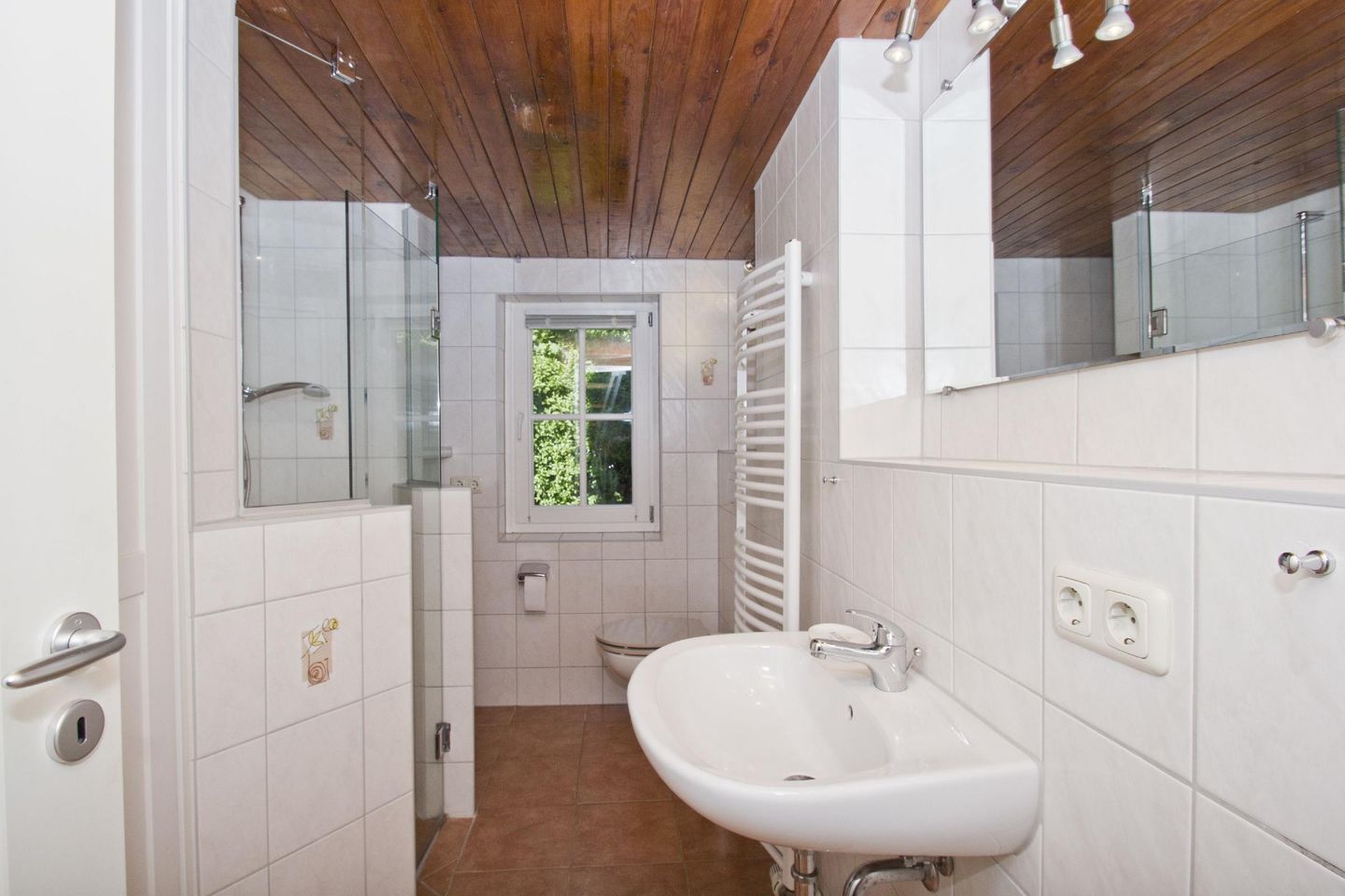 A bathroom with a white sink, a glass shower enclosure, a wooden ceiling, and a window with a view of greenery.